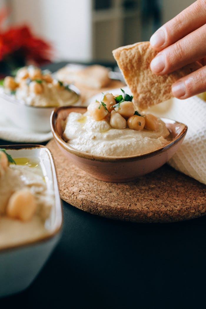about-us-02 Close-up of a hand dipping pita bread into creamy hummus topped with chickpeas.